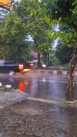 Vehicles drive fast on a wet asphalt road during heavy rain in a tropical suburban area at dusk to demonstrate daily commute challenges and rainy weather conditions.