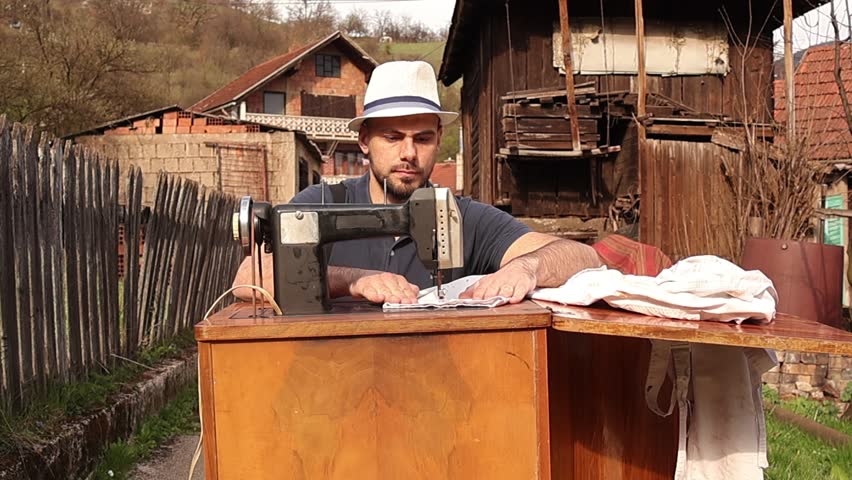 Man in hat and suspenders working at a vintage sewing machine in a rustic setting, slightly leaning forward while closely observing the needle and stitching process, highlighting focus and craftsmanship.