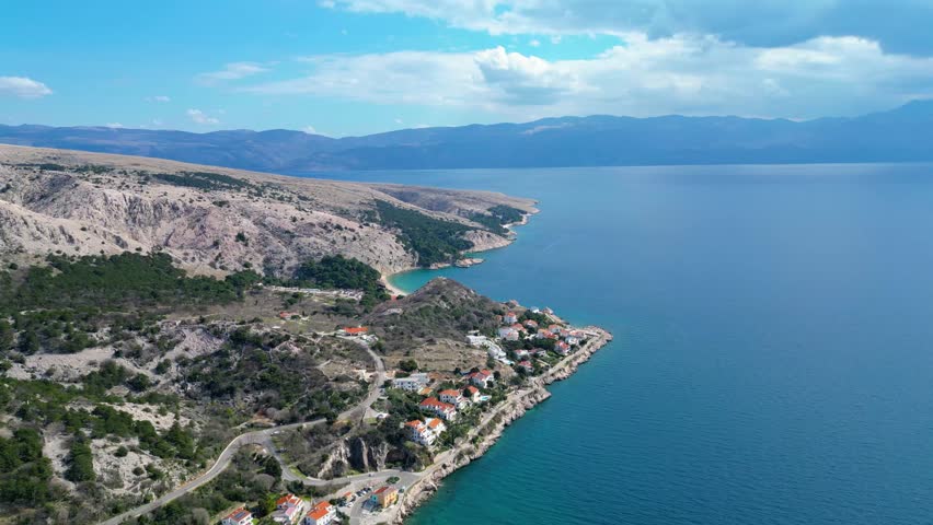 Aerial view of a picturesque Croatian coastal village on Krk Island, with its charming houses nestled along the clear turquoise Adriatic Sea, backed by rugged mountains.
