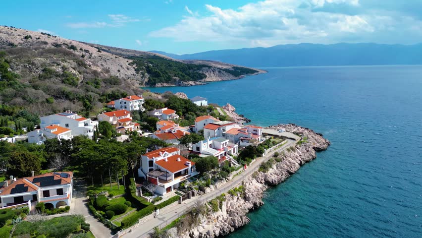 Aerial view of a charming Croatian coastal village on Krk Island, featuring red-roofed homes nestled on rocky cliffs overlooking the clear Adriatic Sea under a bright spring sky.