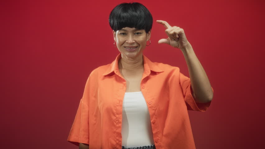 Woman pinching a small gap between thumb and forefinger while smiling in studio with orange shirt visible; playful confidence.