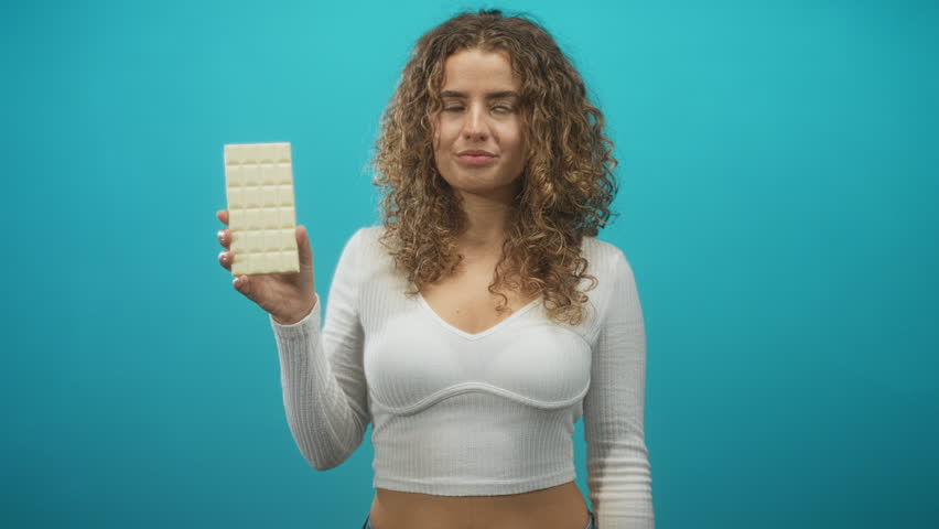Woman holding white chocolate bar with right hand, smiling, curly hair and visible midriff against teal studio backdrop; playful indulgence.