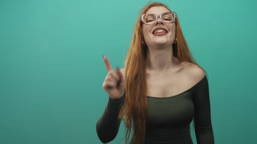 Redhead woman wearing glasses laughing broadly, bare shoulder and collarbone exposed, green off shoulder top against a teal studio backdrop; joy playfulness.
