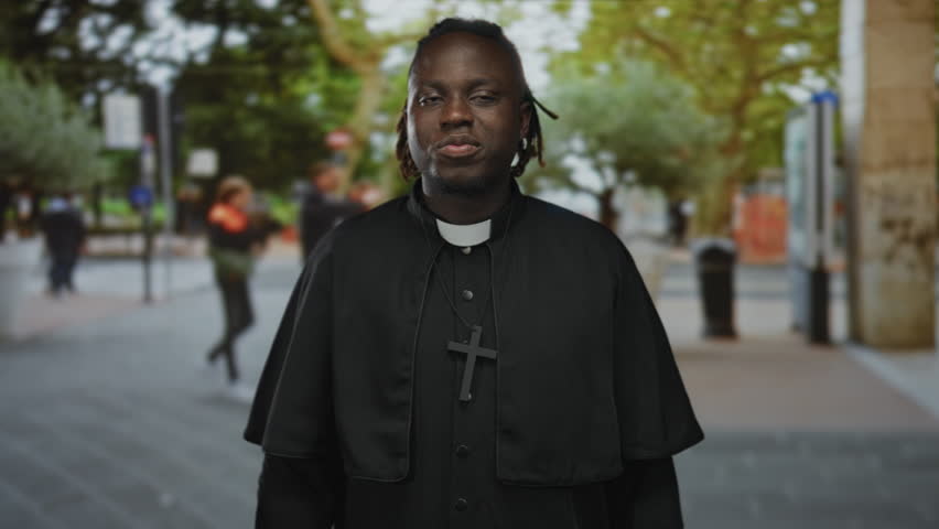 Young man priest wearing clerical collar and cross necklace, finger to lips for silence while standing on a street lined with trees; contemplation.