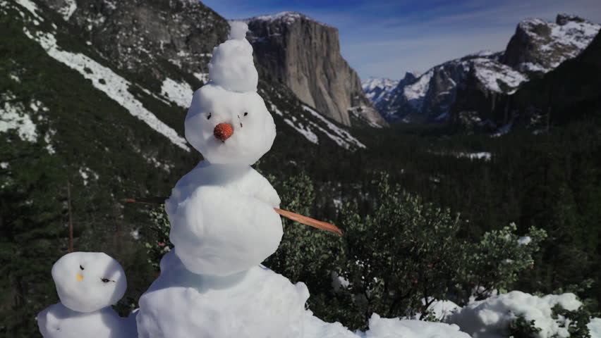 Snowman in yosemite valley winter with El Capitan granite cliffs in background, cheerful handmade snowman and small companion perched on snowy overlook, bright carrot nose and stick arm,