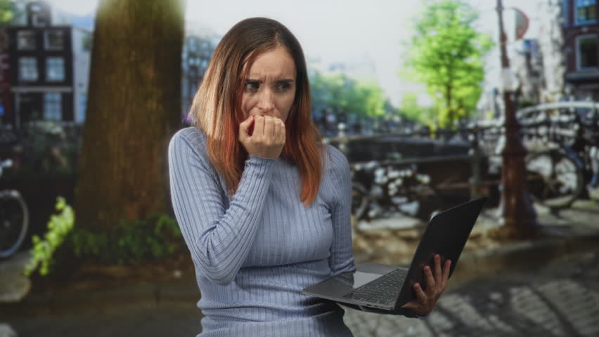 Woman biting nails while holding laptop on a street canal with bicycles and bridge visible; anxiety remote work.