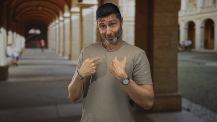 Man points thumbs to chest in building arcade with bracelets and watch visible; awkwardness self reference.