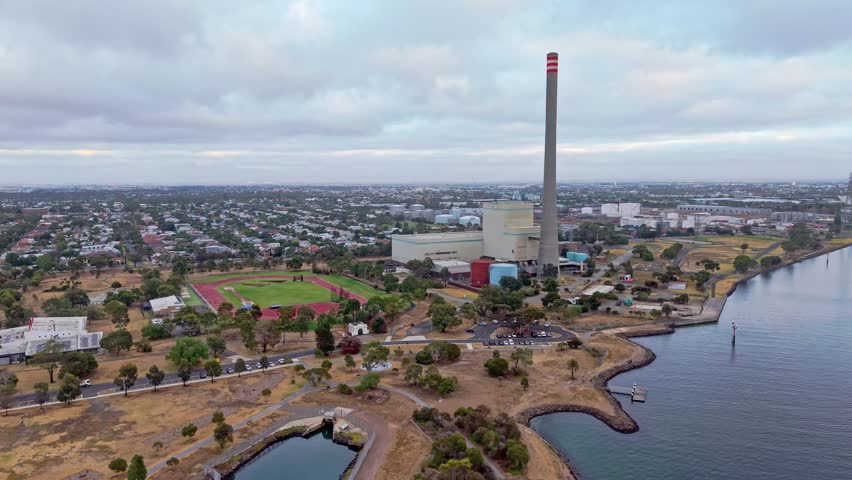 Aerial view of Newport Power Station with its iconic tall chimney beside the waterfront, surrounded by suburban Melbourne. Shows industrial energy infrastructure near residential and coastal areas.