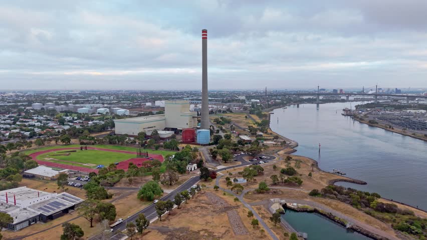 Aerial view of Newport Power Station with its iconic tall chimney beside the waterfront, surrounded by suburban Melbourne. Shows industrial energy infrastructure near residential and coastal areas.