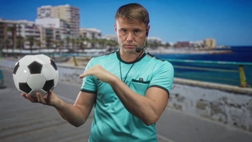 Man holds soccer ball with whistle and headset, palm supporting ball and other hand mid signal while standing on a street with buildings and railing visible; authority confidence.