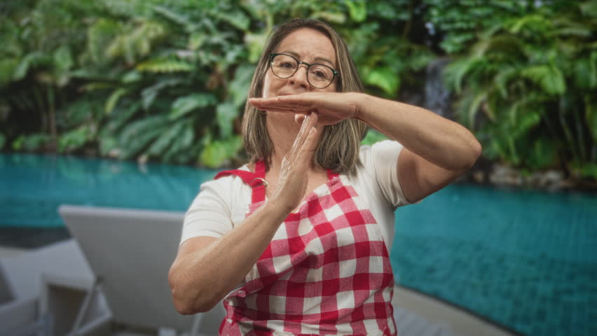 Senior woman in red check apron pouring water between hands while making time out gesture by pool, building; calm focus.
