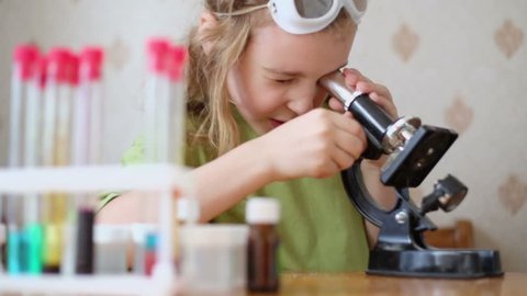 Little girl attentively looks into microscope on table with chemical glassware