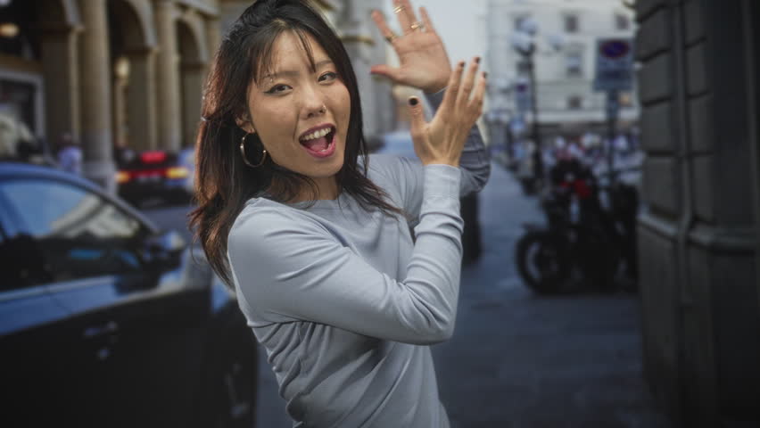 Young asian woman with hoop earrings gesturing with raised hands and open mouth toward parked car and motorcycles on city street; playful curiosity.