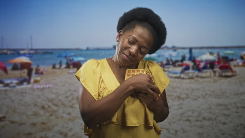 Black woman hugging a waffle to her chest and smiling with closed eyes on a crowded sandy beach with umbrellas and sunbeds; joy comfort nostalgia.