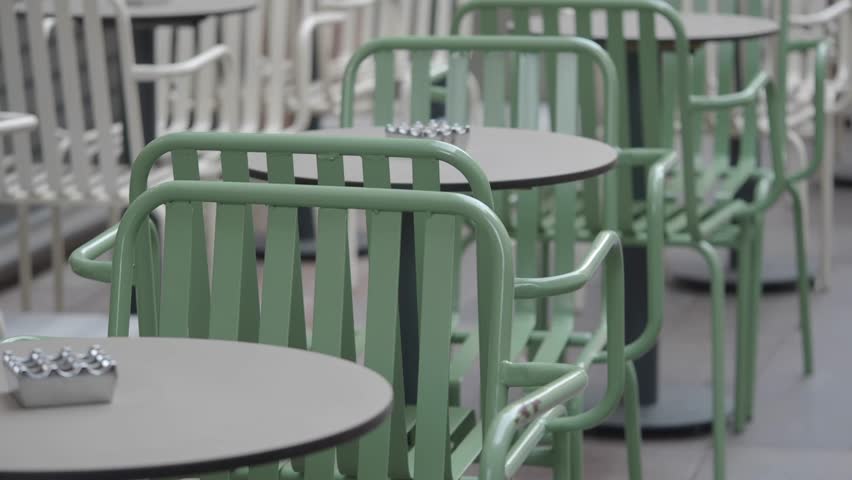 Row of empty tables and chairs at outdoor cafe, green chairs in foreground, white chairs in background, slow motion close up.