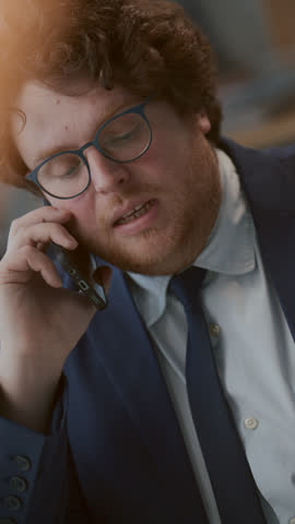 Young businessman in glasses and formal suit holding mobile phone and actively talking during work call in office. Vertical close-up shot