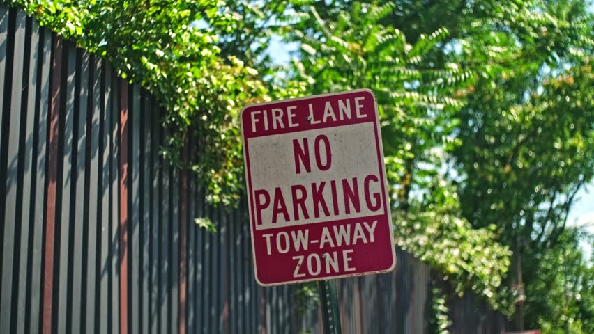 A close up view of an old fire lane no parking tow away zone sign with lush green foliage and a metal fence in background