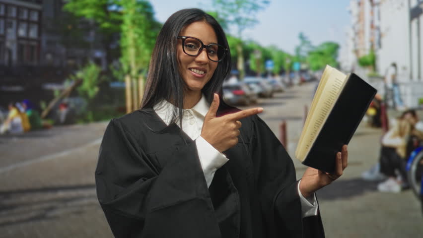 Woman judge in black robe points finger at open book while holding it in hand on a busy street with blurred pedestrians and trees; confidence leadership.