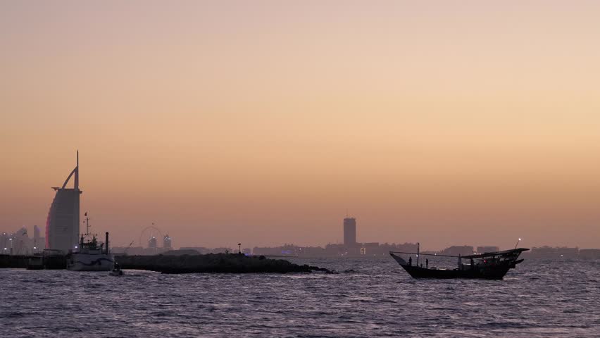 Traditional dhow boat sailing on sea at sunset with dubai cityscape and burj al arab hotel in background at dusk