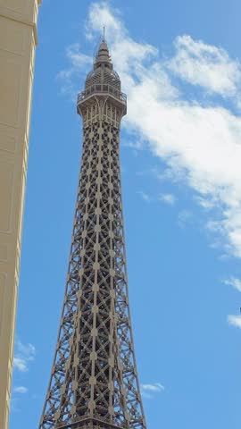 Close-up vertical tilt shot moving from the top to the base of the Eiffel Tower Replica in Las Vegas framed by blue sky, light clouds, palm trees and surrounding high-rise hotel buildings