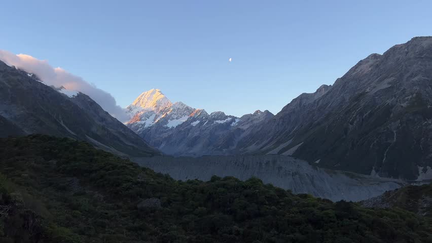 Static shot from Sealy Tarns track looking toward Aoraki Mount Cook at sunset with golden alpenglow on the summit, crescent moon visible, glacier valley below