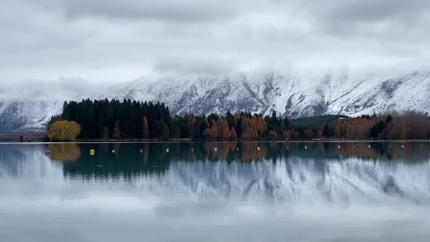 Still lake reflecting snow covered mountain range and autumn colored trees under overcast sky at Lake Ruataniwha, South Island, New Zealand