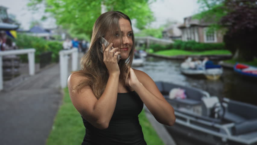 Young woman talking on phone with hand on head on street near canal boats and houses; pensive reflection.