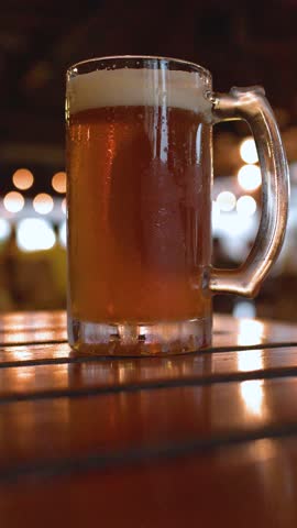 A frosty glass mug of amber beer sits on a wooden table in a bar