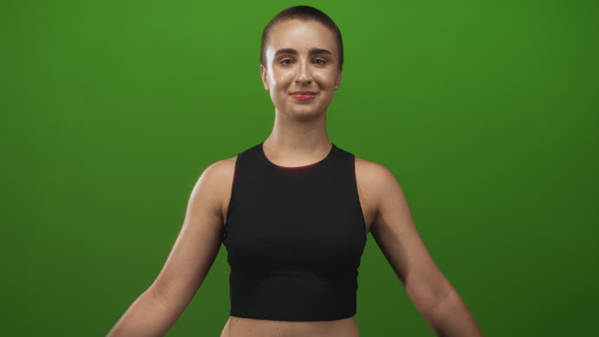 Woman with hands in prayer pose wearing a black tanktop in studio, eyes closed and smiling, green backdrop visible; calm focus.