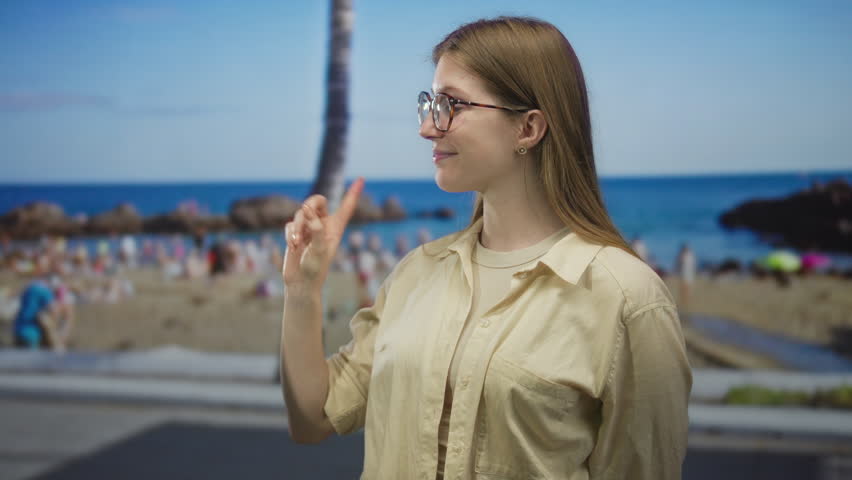 Woman wearing glasses and beige shirt points finger to left on street by palms and crowded beach scene; confidence empowerment determination ambition.