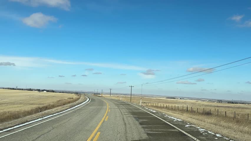 Driving POV, down highway in early spring with open landscape and seasonal transition along rural roadway