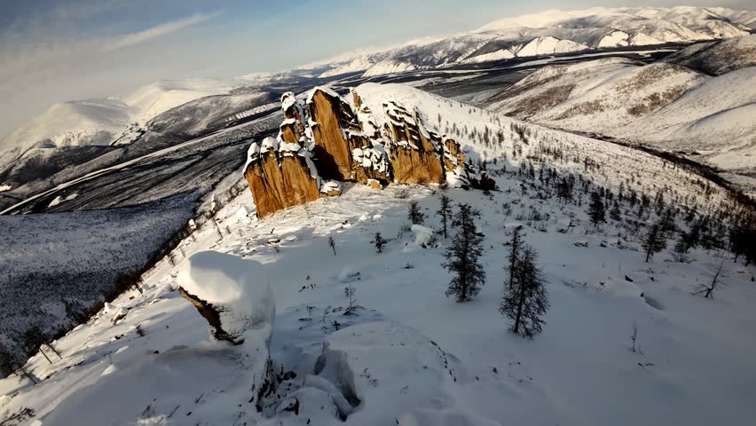 Flying a drone over snowy mountains with rocky cliffs and trees in winter landscape