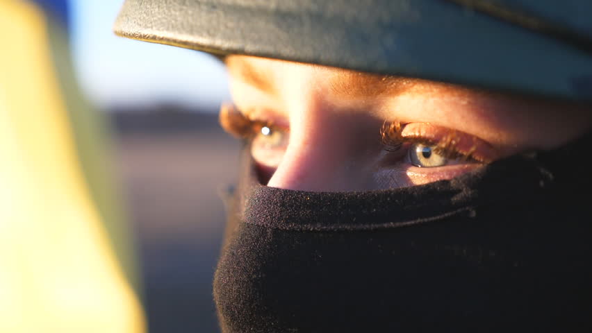 Close up green eyes of female ukrainian army soldier looking at sunset against blue-yellow flag. Sad sight of military woman in helmet and balaclava at countryside. Resistance to russian invasion