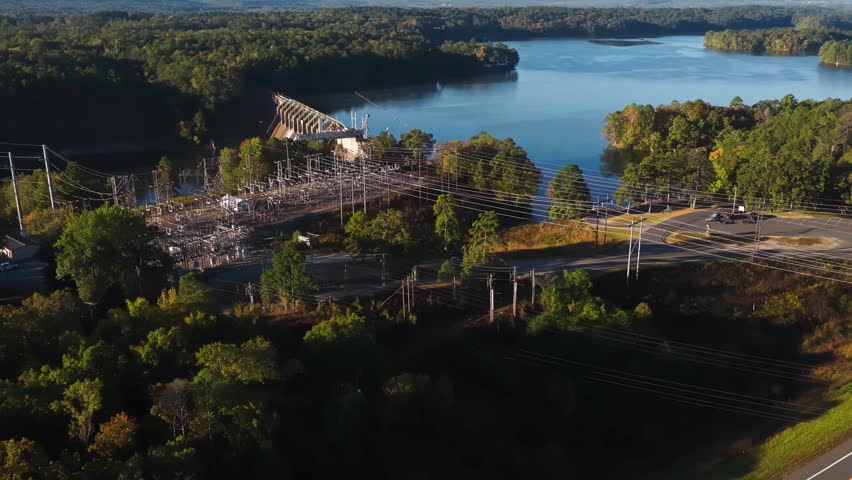 Electrical substation near reservoir and hydroelectric dam surrounded by forested hills, renewable energy infrastructure in Arkansas USA, aerial reveal