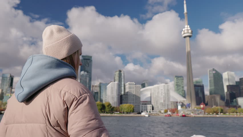 Young woman tourist looking at Toronto skyline from waterfront, back view of teenager in warm clothes enjoying cityscape and travel atmosphere on Lake Ontario.