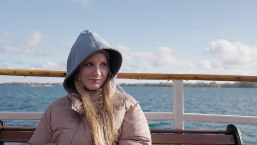 Teenage girl passenger sitting on boat bench on Lake Ontario, young woman in hooded jacket enjoying calm water travel.
