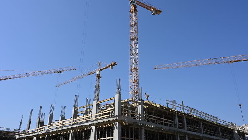 Several yellow tower cranes operate above a building under construction featuring concrete pillars and steel reinforcement. The scene shows workers on the top level against a clear blue sky.