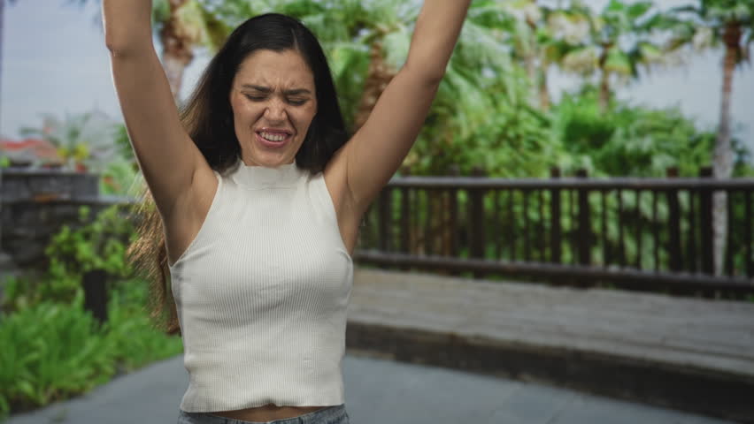 Woman waving hands in park with raised arms and shouting while calling for someone on a green outdoor walkway; calling excitement.