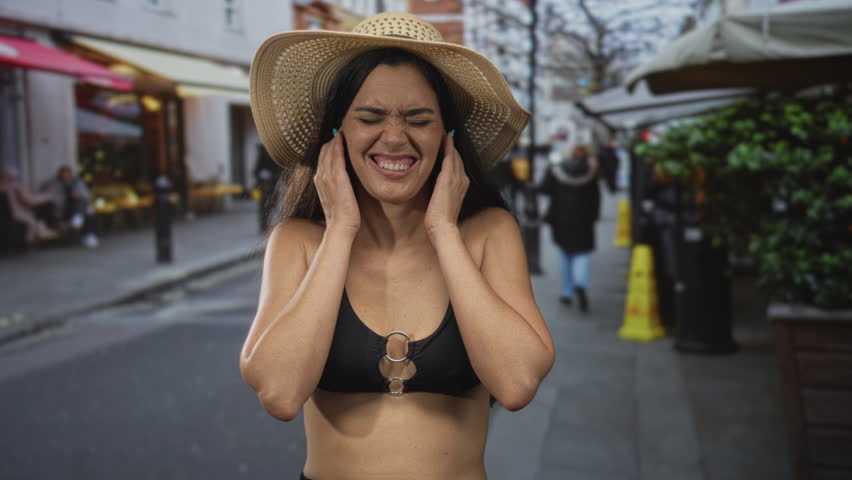 Woman wearing straw hat and black bikini top covers ears and winces on busy street amid sidewalk cafe tables and pedestrians; stress.