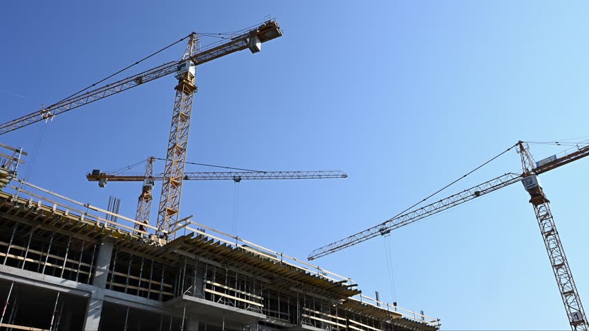 An active construction site featuring several yellow tower cranes operating above a reinforced concrete building frame. The scene captures structural development, metal shoring props, and workers against a clear blue sky.