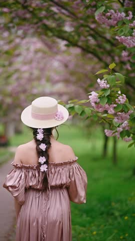 Young woman in pink dress standing under blooming sakura tree, holding straw hat and decorated braid with sakura flowers, romantic spring portrait