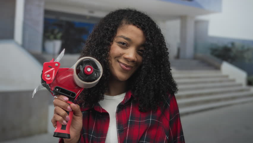 African american woman holds red packing tape dispenser on building steps with bright smile; joy.