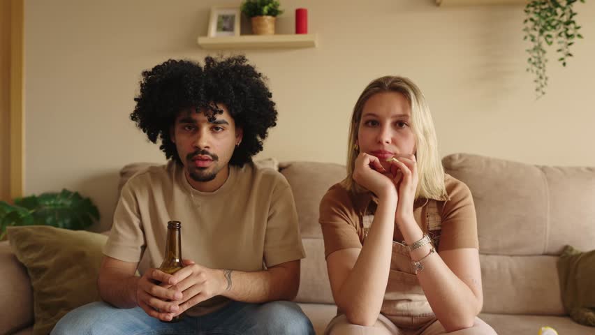 Anxious interracial couple sitting on a sofa and watching a sports match. Young man and woman looking sad after their favorite team loses