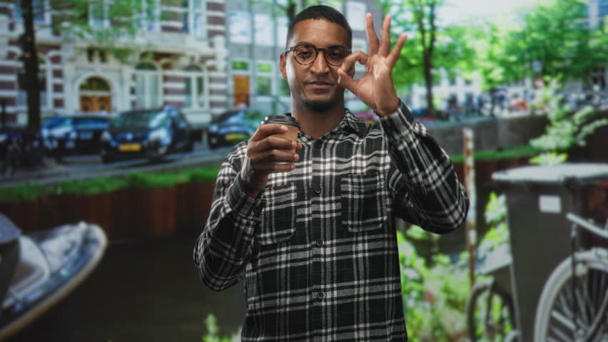 Young black man in plaid shirt and glasses holding takeaway coffee cup while making ok sign to eye on street with canal boats behind; playful curiosity charm.