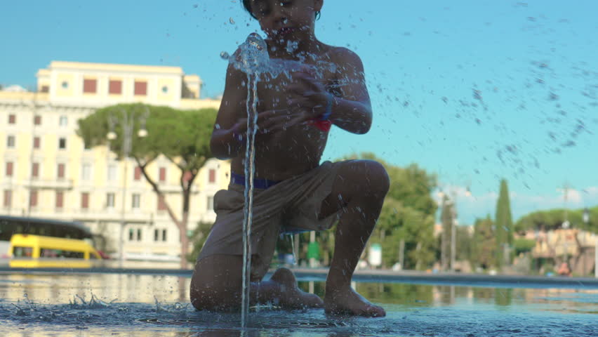 Boy playfully fighting water fountain jet with punches and kicks on wet pavement in public square during summer day creating energetic and carefree scene
