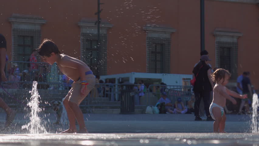 Older boy and toddler playing near fountain streams, carefree children splashing water together in public square, joyful summer innocence and playful energy