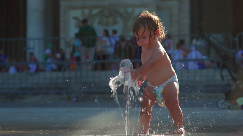 Toddler standing next to fountain stream, water splashing on feet while cooling off and playing joyfully on summer day outdoors