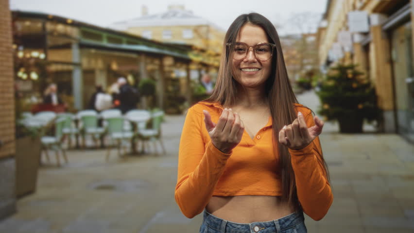 Woman showing hands palms up, wearing glasses and orange crop top on a street lined with cafe tables and a decorated tree, smiling and relaxed; welcoming warmth joy.