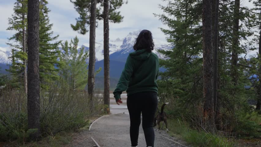 Woman and dog walking on a trail with snowy mountains and pine trees in Jasper National Park