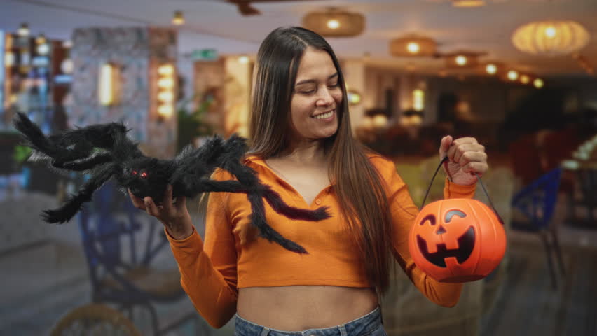 Woman holds pumpkin bucket and large black spider with exposed midriff and smiling expression in a restaurant building; playful celebration.